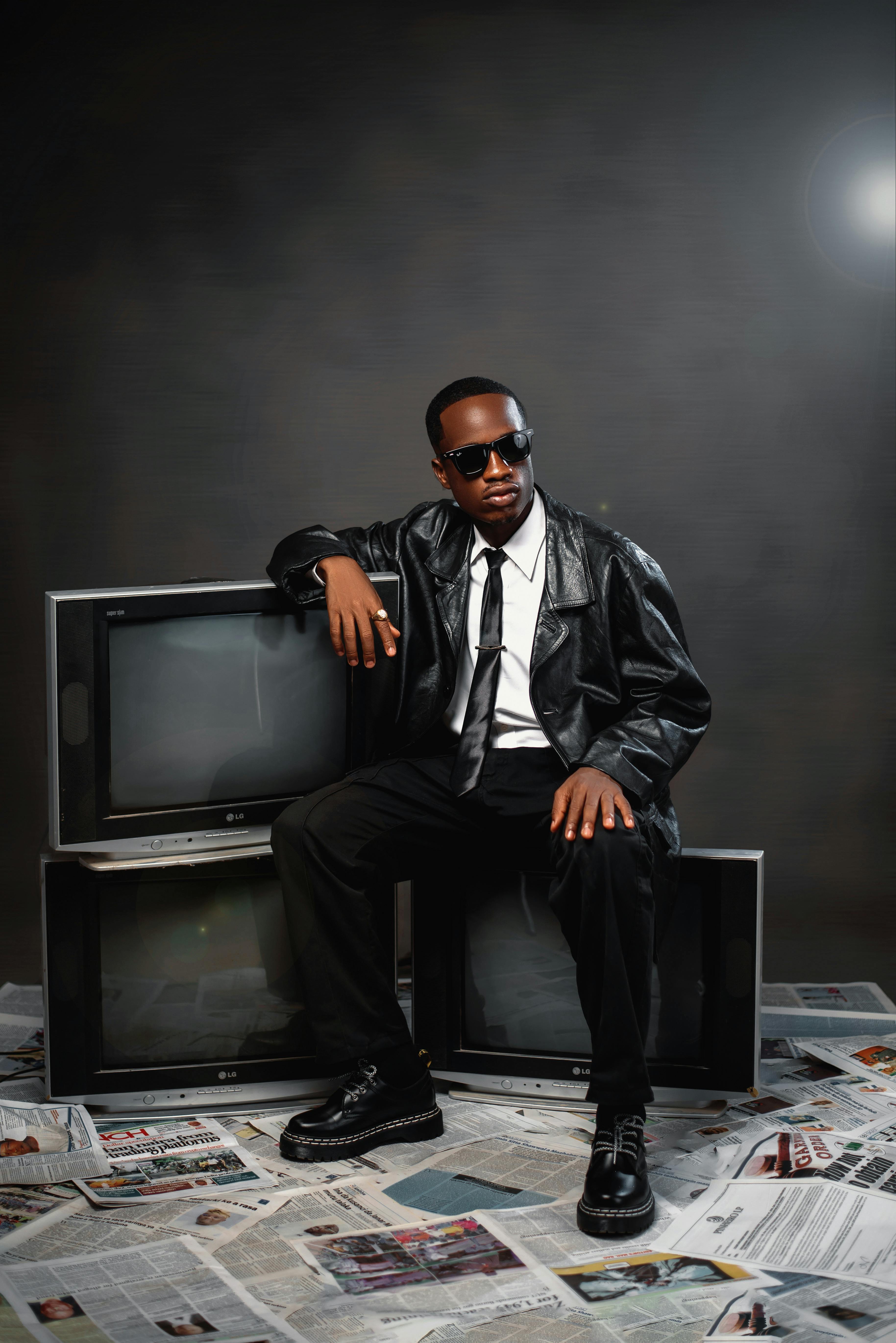 Person in leather jacket and sunglasses sits on retro TVs surrounded by newspapers.